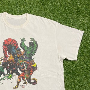 Op de afbeelding: Wit T-shirt met een kleurrijke grafische print van verschillende superhelden die tegen Thanos vechten. De grafiek bevat Hulk, The Thing en andere personages. Het shirt heeft een ronde hals en korte mouwen.