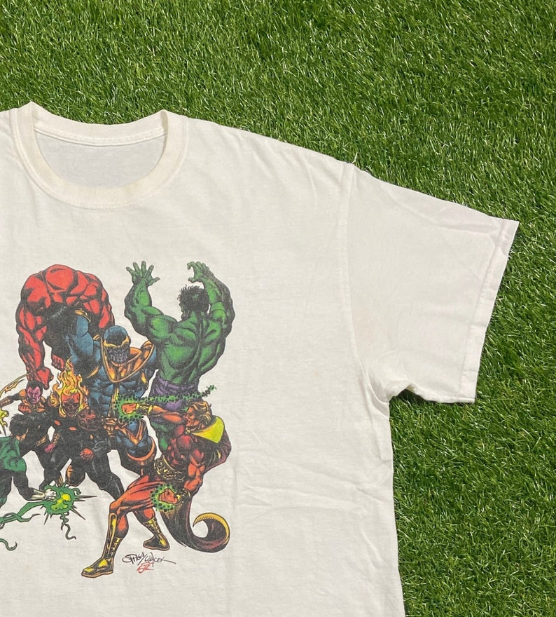 Op de afbeelding: Wit T-shirt met een kleurrijke grafische print van verschillende superhelden die tegen Thanos vechten. De grafiek bevat Hulk, The Thing en andere personages. Het shirt heeft een ronde hals en korte mouwen.