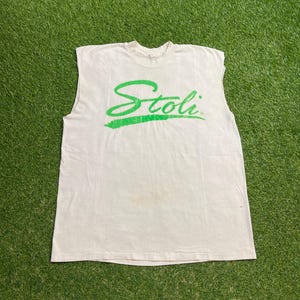 以下が含まれることがあります： 胸に緑色の筆記体で「Stoli」とプリントされた白いノースリーブTシャツ。テキストは筆記体で書かれています。この服はヴィンテージのようで、シミがあります。