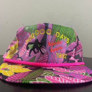 Puede incluir: Una gorra de béisbol de color rosa, negro y verde con estampado floral y ribete de cuerda rosa. La parte delantera de la gorra tiene bordado el texto "Hoo-Doo Days, Neola Iowa 1992" con un gráfico de gato negro.