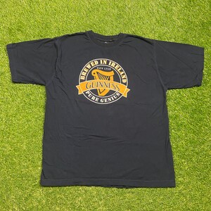 May include: A dark blue t-shirt with a gold circle graphic featuring a harp, the text "Brewed in Ireland Est 1759 Guinness Pure Genius", and a gold ribbon.