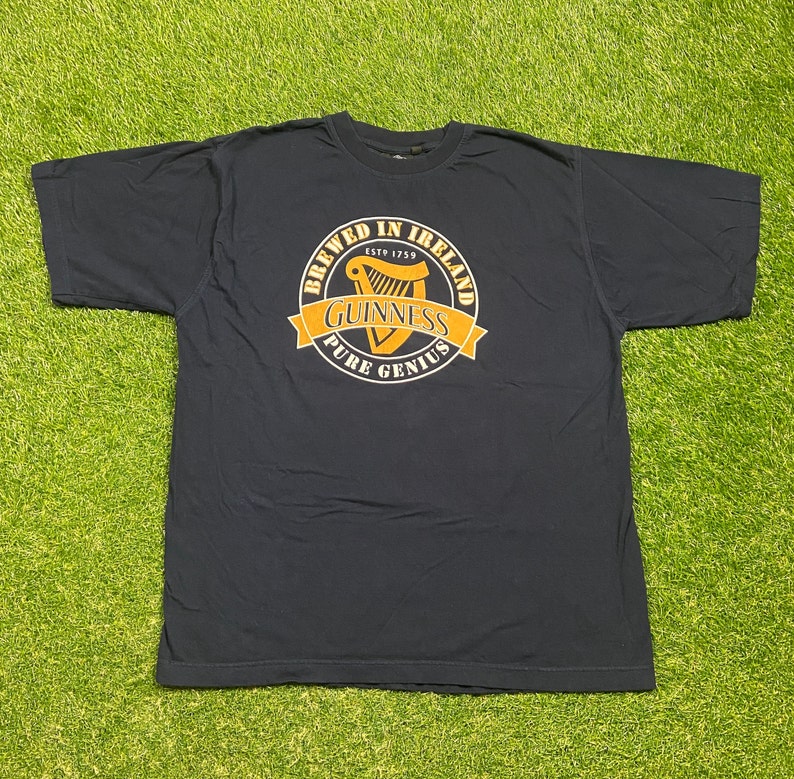 May include: A dark blue t-shirt with a gold circle graphic featuring a harp, the text "Brewed in Ireland Est 1759 Guinness Pure Genius", and a gold ribbon.