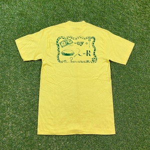 Peut inclure: T-shirt jaune à manches courtes avec un motif vert représentant une tarte, de l'argent, un club de golf et le texte "Hey" et "R".