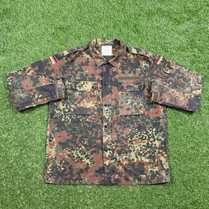 Puede incluir: Una camisa de botones de camuflaje con mangas largas y dos bolsillos en el pecho. La camisa tiene un parche de la bandera alemana en la manga izquierda y un pequeño parche en la manga derecha.