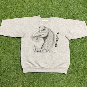 Op de afbeelding: Een lichtgrijs sweatshirt met een zwart-wit illustratie van een Brachiosaurus dinosaurus die bladeren eet, met het woord "Brachiosaurus" verticaal gedrukt. Er zijn ook verschillende kleinere dinosaurussen afgebeeld.