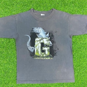 Può includere: Una maglietta grigia scura con una stampa grafica della testa di Godzilla che emerge da una lettera G. Il testo "Godzilla" è stampato sotto la grafica.