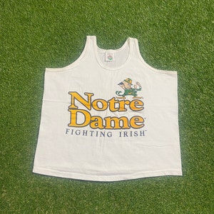 Puede incluir: Camiseta sin mangas blanca con un logotipo amarillo y verde de Notre Dame Fighting Irish. El logotipo presenta un duende de dibujos animados.