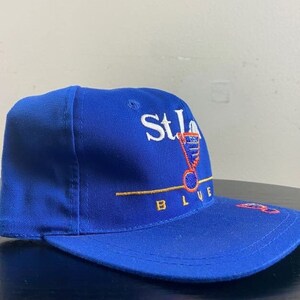 K&ouml;nnte beinhalten: Eine blaue Baseballkappe mit dem gestickten Logo der St. Louis Blues auf der Vorderseite. Das Logo ist wei&szlig; mit roten und goldenen Akzenten. Der Text "St. Louis Blues" ist in Wei&szlig; unter dem Logo gestickt.