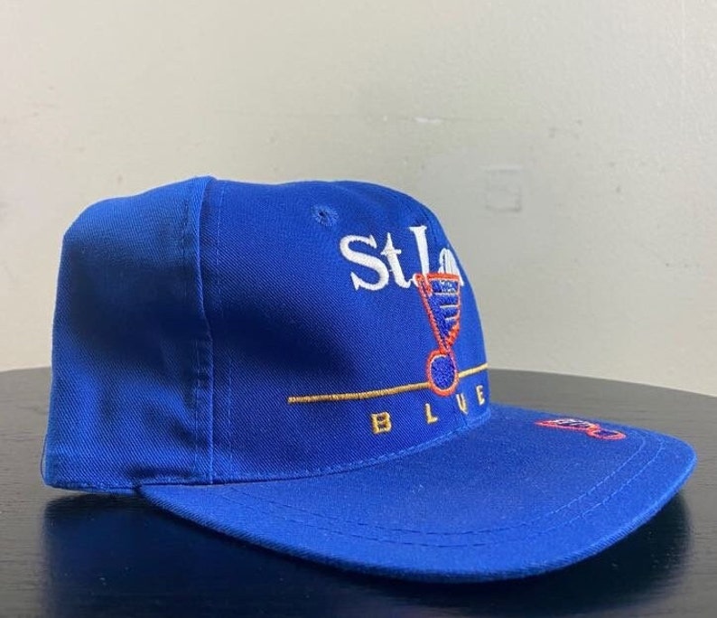 K&ouml;nnte beinhalten: Eine blaue Baseballkappe mit dem gestickten Logo der St. Louis Blues auf der Vorderseite. Das Logo ist wei&szlig; mit roten und goldenen Akzenten. Der Text "St. Louis Blues" ist in Wei&szlig; unter dem Logo gestickt.
