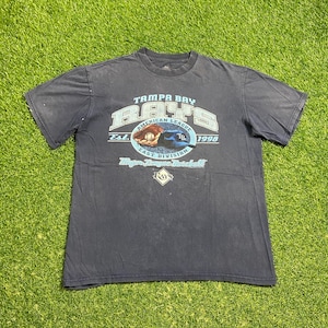 May include: A navy blue t-shirt featuring the Tampa Bay Rays logo. The design includes the team name, "American League East Division," and the year "1998." The shirt has a crew neck and short sleeves, with a faded appearance.