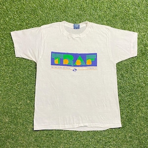 Puede incluir: Camiseta blanca con un diseño gráfico con las letras "TRAF" en verde y naranja, sobre un fondo rectangular azul y morado. Debajo del gráfico está el texto que dice "1988 Three Rivers Arts Festival Pittsburgh, PA."
