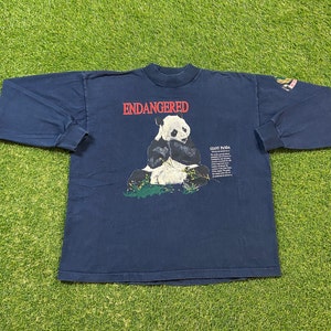 May include: A navy blue long-sleeved t-shirt with a graphic of a giant panda sitting on a bed of green leaves. The text "ENDANGERED" is printed in red above the panda. The text "GIANT PANDA" is printed in black below the panda.