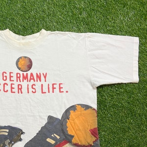 May include: White t-shirt with a red and black graphic print of a soccer ball with the text "GERMANY SOCCER IS LIFE."
