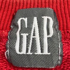 Pu&ograve; includere: Una felpa rossa con una toppa grigia che dice "GAP" in lettere bianche.