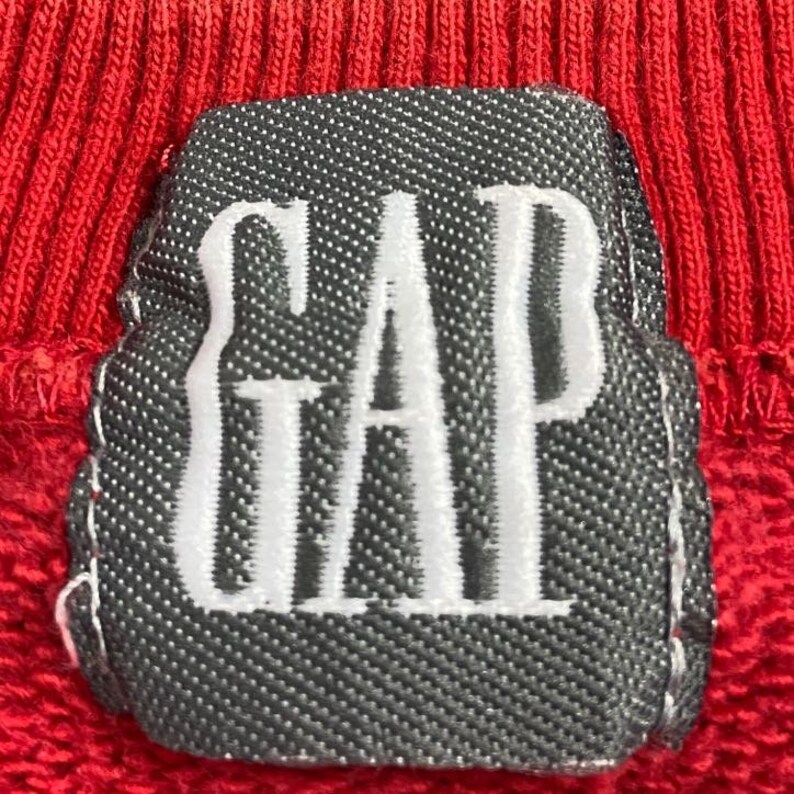 Pu&ograve; includere: Una felpa rossa con una toppa grigia che dice "GAP" in lettere bianche.