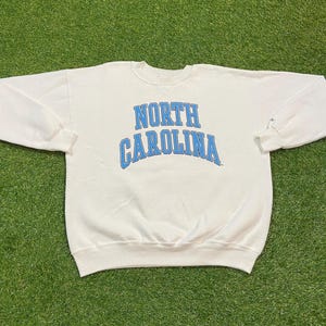 May include: A white crewneck sweatshirt with the words "NORTH CAROLINA" in light blue arched lettering. The sweatshirt has long sleeves and a ribbed neckline and cuffs. The garment is laid out on a green surface.