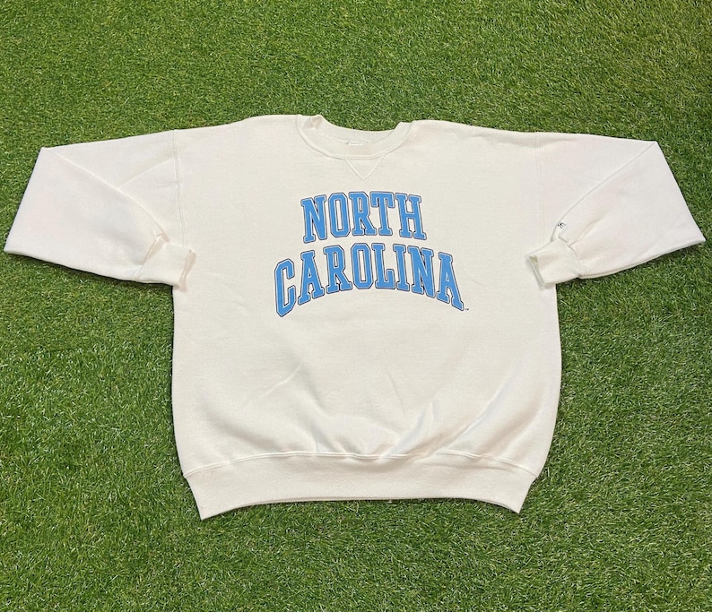 May include: A white crewneck sweatshirt with the words "NORTH CAROLINA" in light blue arched lettering. The sweatshirt has long sleeves and a ribbed neckline and cuffs. The garment is laid out on a green surface.