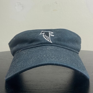 May include: A black visor with a white embroidered Atlanta Falcons logo. The logo is a stylized bird with wings spread.