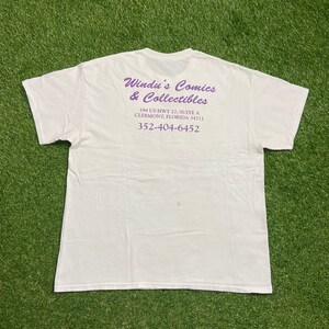 Op de afbeelding: Wit T-shirt met paarse tekst die "Windu's Comics & Collectibles",  "194 US HWY 27, SUITE A", "CLERMONT, FLORIDA 34711" en "352-404-6452" luidt.