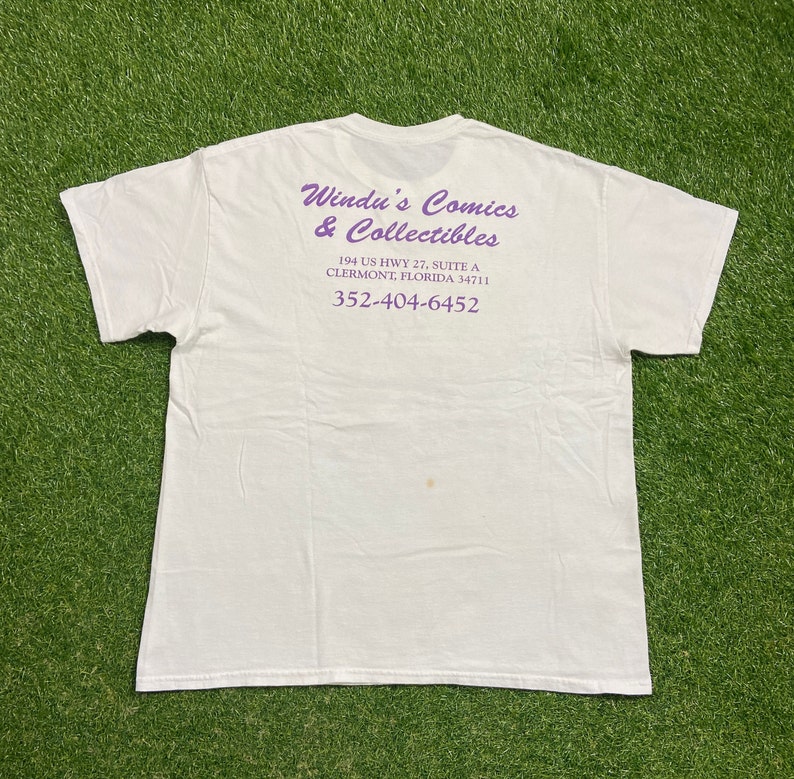 Op de afbeelding: Wit T-shirt met paarse tekst die "Windu's Comics & Collectibles",  "194 US HWY 27, SUITE A", "CLERMONT, FLORIDA 34711" en "352-404-6452" luidt.