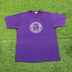 May include: Purple t-shirt featuring a white circular graphic with the text "Tarleton State University Stephenville, Texas 1899" surrounding a "T" shape. The shirt is made of a soft material and has short sleeves.