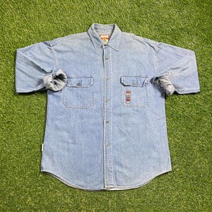 May include: A light blue denim shirt with long sleeves, a button-down front, and two chest pockets. The shirt has a small embroidered patch on the left chest pocket.