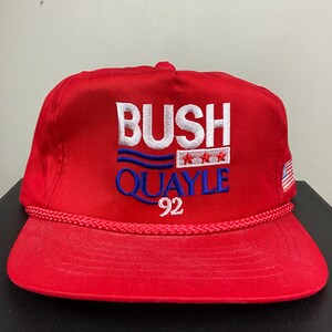 Può includere: Cappellino da baseball rosso con la scritta ricamata "BUSH QUAYLE 92" in bianco e blu. Il cappellino presenta un dettaglio a corda lungo il bordo e una piccola toppa con la bandiera americana sul lato.