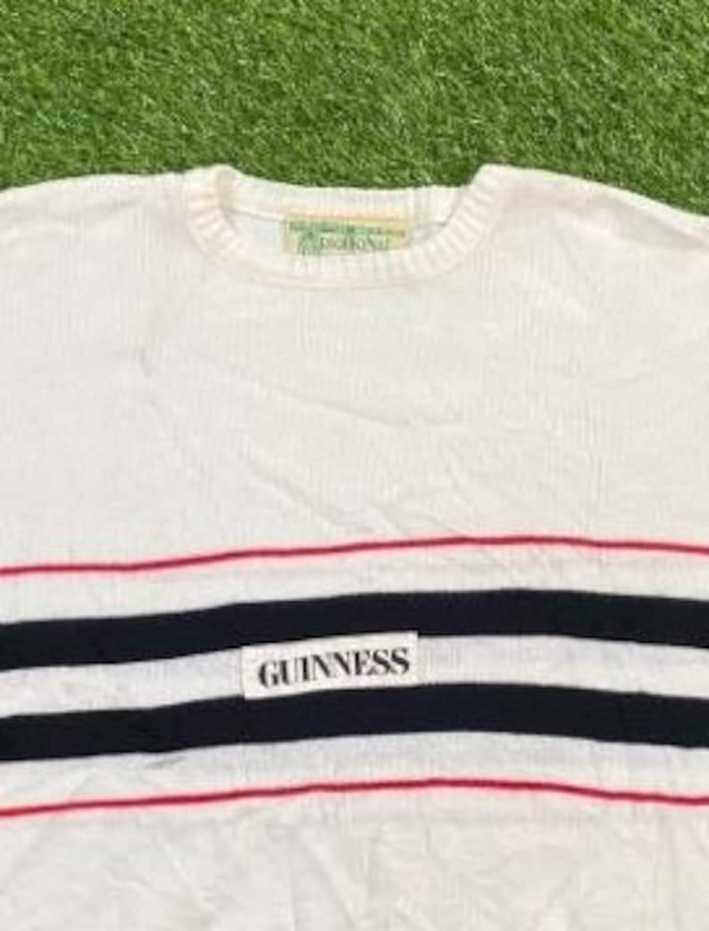 May include: White long-sleeve sweater with red and black stripes and a white label with the text "GUINNESS" in black.