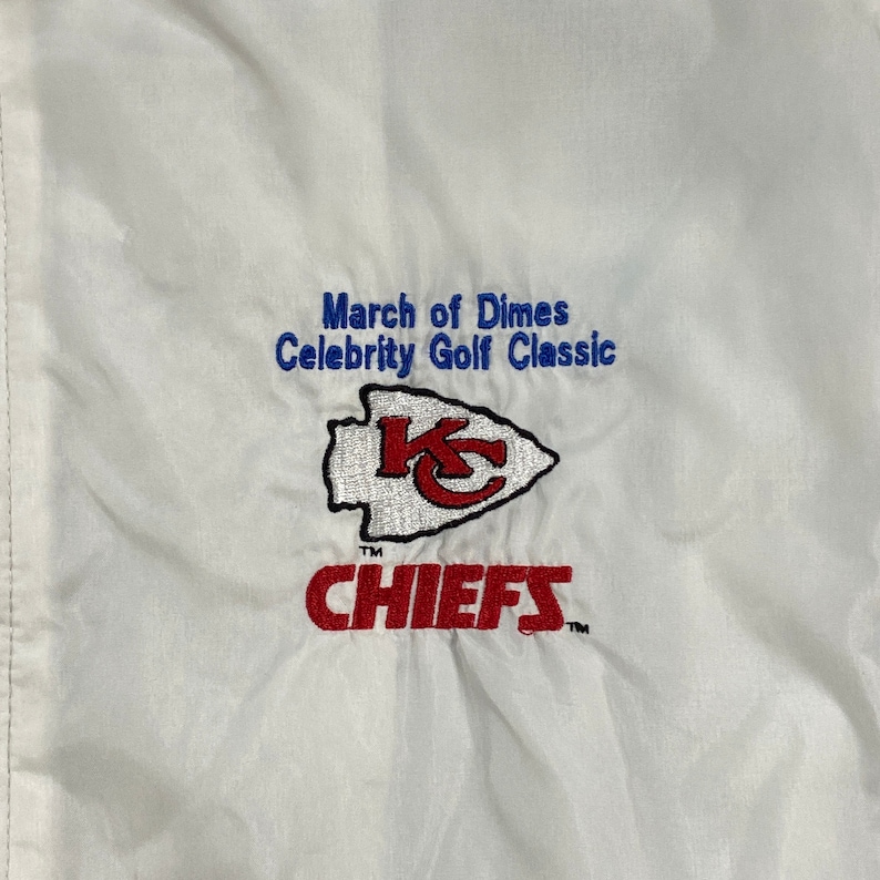 May include: White windbreaker jacket with a red and white Kansas City Chiefs logo and the text "March of Dimes Celebrity Golf Classic" in blue.