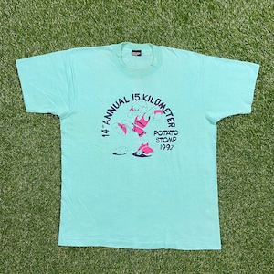 Pode incluir: Uma camiseta azul clara com um gráfico de um personagem de desenho animado correndo e o texto "14th Annual 15 Kilometer Potato Stomp 1992".