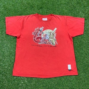 May include: Red t-shirt with a graphic print of a cartoon character holding a baseball bat and a woman in a green dress. The text "Land of the Free... Land of the Crooked." is printed below the graphic.