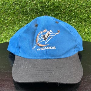 Puede incluir: Gorra de béisbol azul y negra con el logotipo de los Wizards bordado en la parte delantera. La gorra tiene una visera curva y un diseño de seis paneles. El logotipo incluye una pelota de baloncesto, una estrella y la palabra "Wizards".