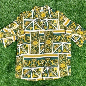 May include: A button-down shirt with a repeating pattern of gold, white, and green geometric shapes and floral designs. The short-sleeved shirt is made of a lightweight fabric and is laid out on a green surface.