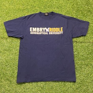 May include: Navy blue t-shirt with the text "EMBRY RIDDLE AERONAUTICAL UNIVERSITY" in white and gold. The shirt is laid flat on a green surface. The t-shirt is a crew neck style.