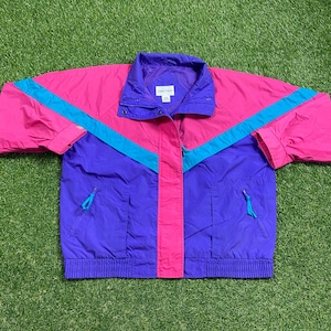 May include: A pink, purple, and turquoise color block windbreaker jacket with a zipper closure and two pockets. The jacket has a small white label with the text "Camp Gear" on the front.
