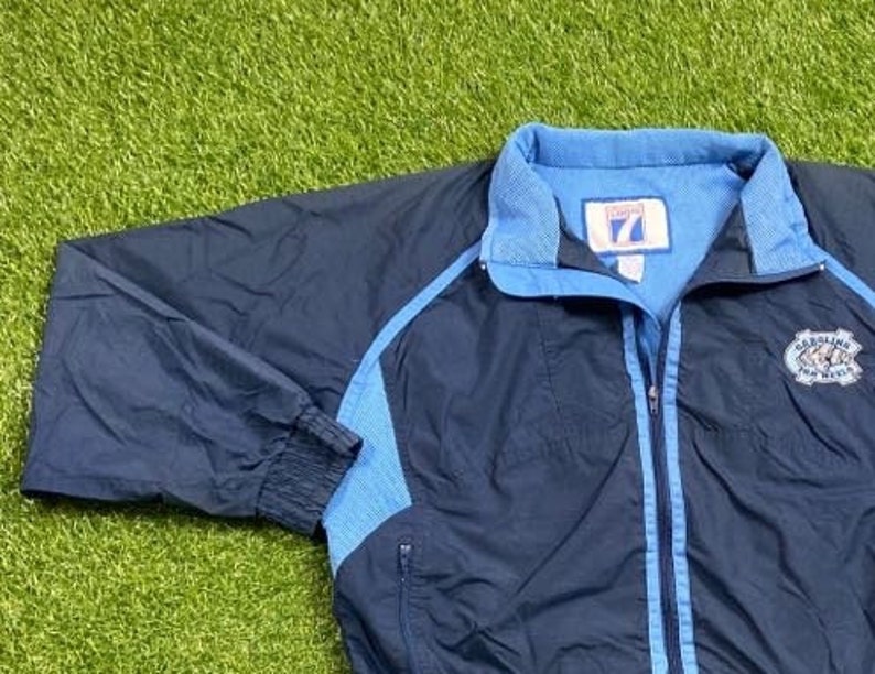 Peut inclure: Une veste coupe-vent bleu marine avec des accents bleu clair et un logo Carolina Tar Heels sur le c&ocirc;t&eacute; droit. La veste a une fermeture &eacute;clair et deux poches.