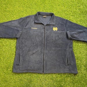Puede incluir: Chaqueta polar Columbia azul marino con cremallera completa, dos bolsillos y el logotipo de Notre Dame en dorado en el pecho. El logotipo de Columbia está bordado en dorado en el pecho izquierdo.