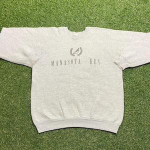Puede incluir: Una sudadera gris claro con mangas largas. El texto "MANASOTA KEY" está impreso en un color gris más oscuro en el pecho, con un diseño de velero y corona encima del texto. La sudadera está hecha de un material suave.
