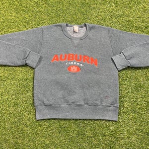 May include: A gray crewneck sweatshirt with the word "AUBURN" in orange block letters across the chest, with "TIGERS" below and a logo. The sweatshirt has long sleeves and a ribbed hem and cuffs. The sweatshirt is lying on a green surface.