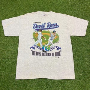 May include: A light gray t-shirt featuring the Tampa Bay Devil Rays logo with baseball players and the text "The Boys Are Back In Town." The shirt has a crew neck and short sleeves.