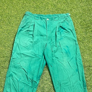 Op de afbeelding: Felgroene geplooide broek. De broek heeft een knoopsluiting en zakken. De stof lijkt een stevig katoen of een mix te zijn. De broek ligt op een groene, grasachtige ondergrond.