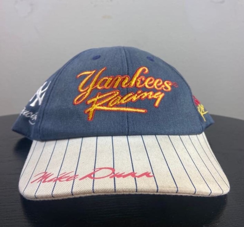 May include: A navy blue baseball cap with "Yankees Racing" embroidered in yellow and red script. The brim is white with navy pinstripes and the name "Mike Dunn" in red script. The cap features the Yankees logo.