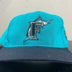 May include: A teal blue baseball cap with a black brim. The front of the cap features a black and white embroidered logo of a marlin fish with the letter "F" in the middle.