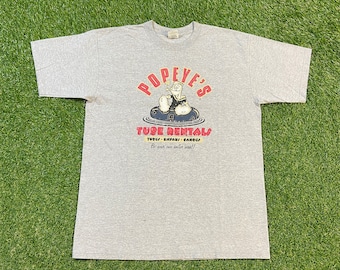 May include: A gray t-shirt with a cartoon image of Popeye the Sailor Man on a tube. The text "Popeye's Tube Rentals" is printed in red and black with the words "Tubes - Kayaks - Canoes" and "Be your own sailor man!!" printed below.