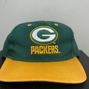 Puede incluir: Gorra de béisbol verde y dorada con el logotipo de los Green Bay Packers y la palabra "Packers" bordada en la parte delantera.