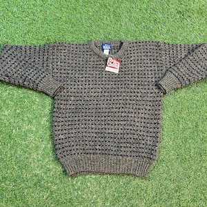 Puede incluir: Un suéter de punto marrón y negro con un patrón texturizado. El suéter tiene cuello redondo y mangas largas. El suéter tiene una pequeña etiqueta en la parte delantera que dice "Woolrich".