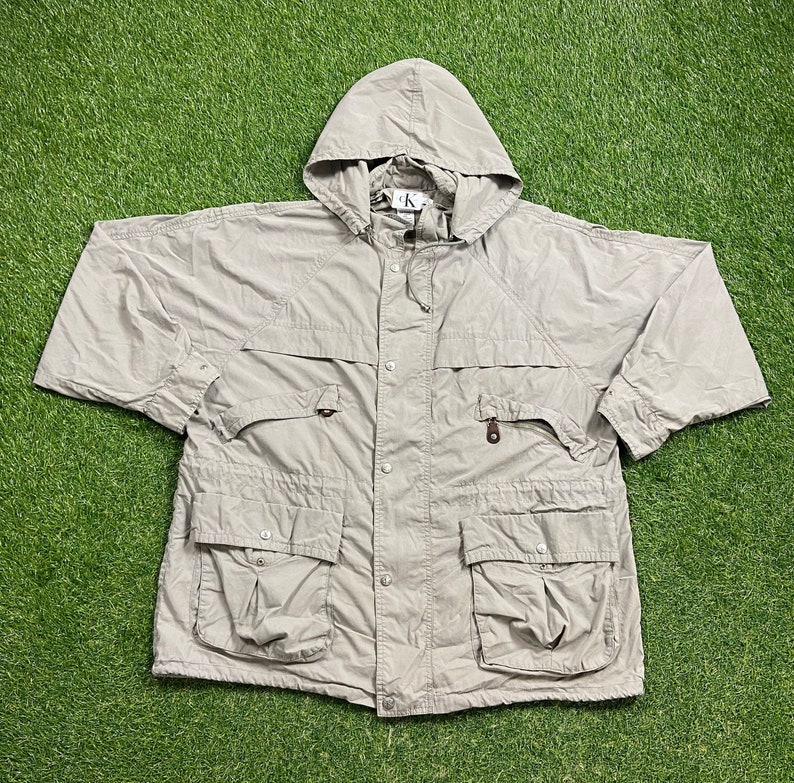 Pu&ograve; includere: Giacca con cappuccio beige chiaro con pi&ugrave; tasche e chiusura con bottoni. La giacca ha un cappuccio con coulisse e un piccolo logo vicino al colletto. Realizzata in materiale leggero, ideale per la stratificazione. Su sfondo verde.