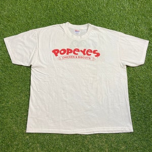 Könnte beinhalten: Weißes T-Shirt mit dem roten Logo "Popeyes Chicken & Biscuits" auf der Brust. Das Shirt besteht aus weichem Material und hat kurze Ärmel. Das T-Shirt liegt auf einer grünen Oberfläche.