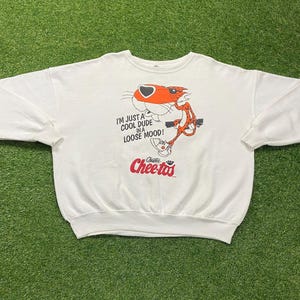 Pode incluir: Camisola branca de gola redonda com um gráfico de Chester Cheetah, a mascote dos Cheetos, e o texto "I'm just a cool dude in a loose mood!". O logótipo dos Cheetos também é visível. O gráfico é em laranja, preto e branco.