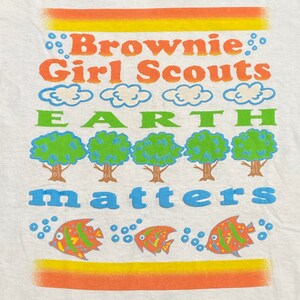 K&ouml;nnte beinhalten: Wei&szlig;es T-Shirt mit einem bunten Grafikdesign. Das Design zeigt den Text "Brownie Girl Scouts" und "Earth Matters" mit einer Reihe von B&auml;umen und Fischen.